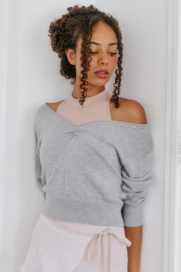 Woman wearing a gray off-shoulder sweater and white skirt against a white wall.